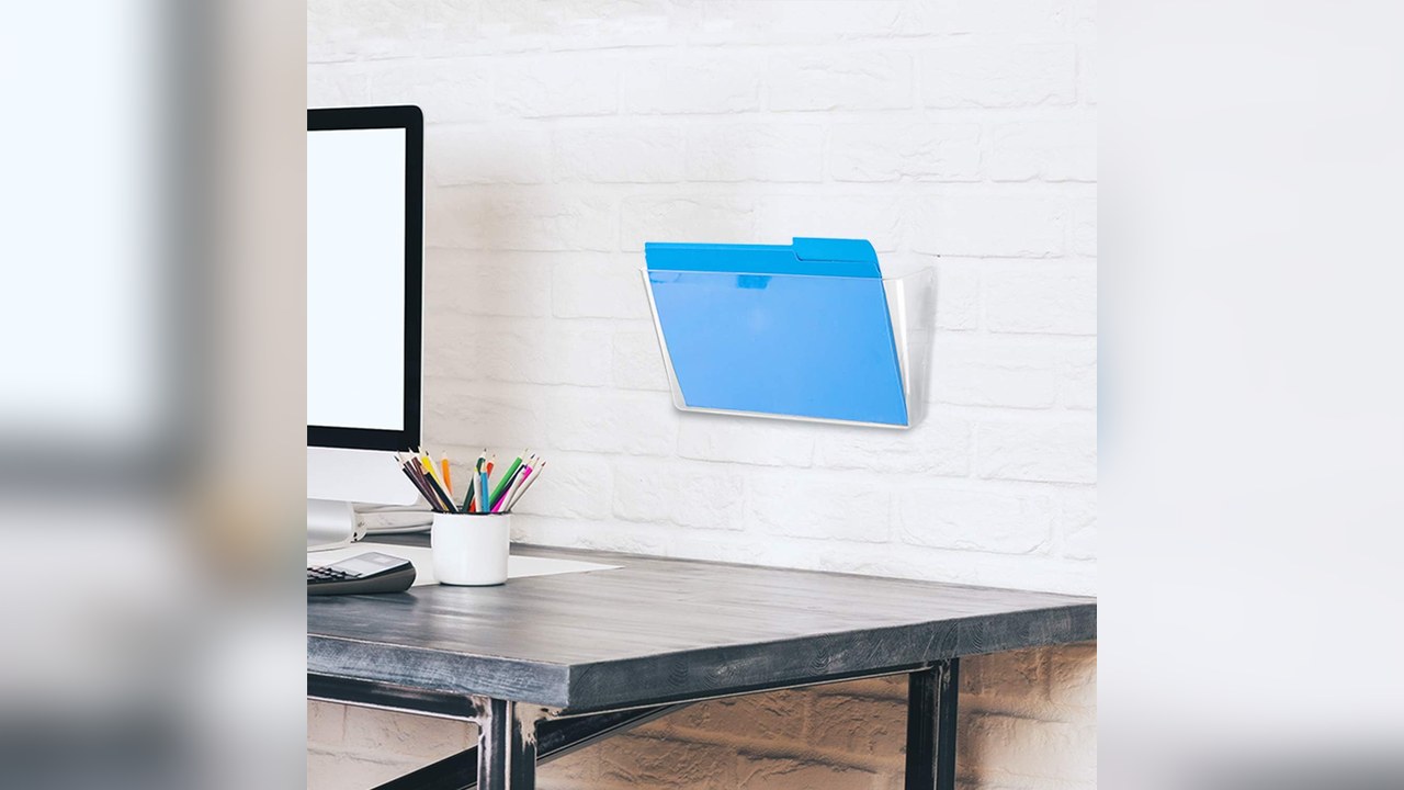 Amazon Basics clear plastic wall file holder with blue folder mounted on white brick wall above desk