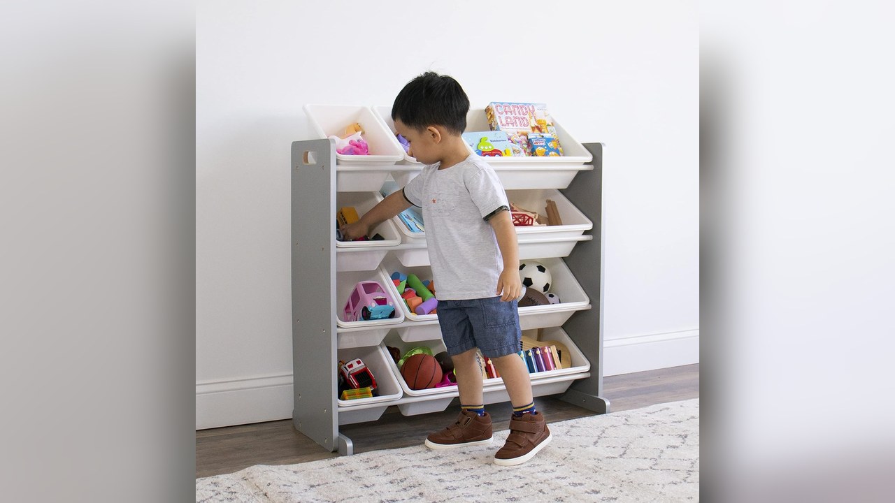 Grey and white four tier toy organizer with twelve plastic bins in a living room while a toddler picks out a toy