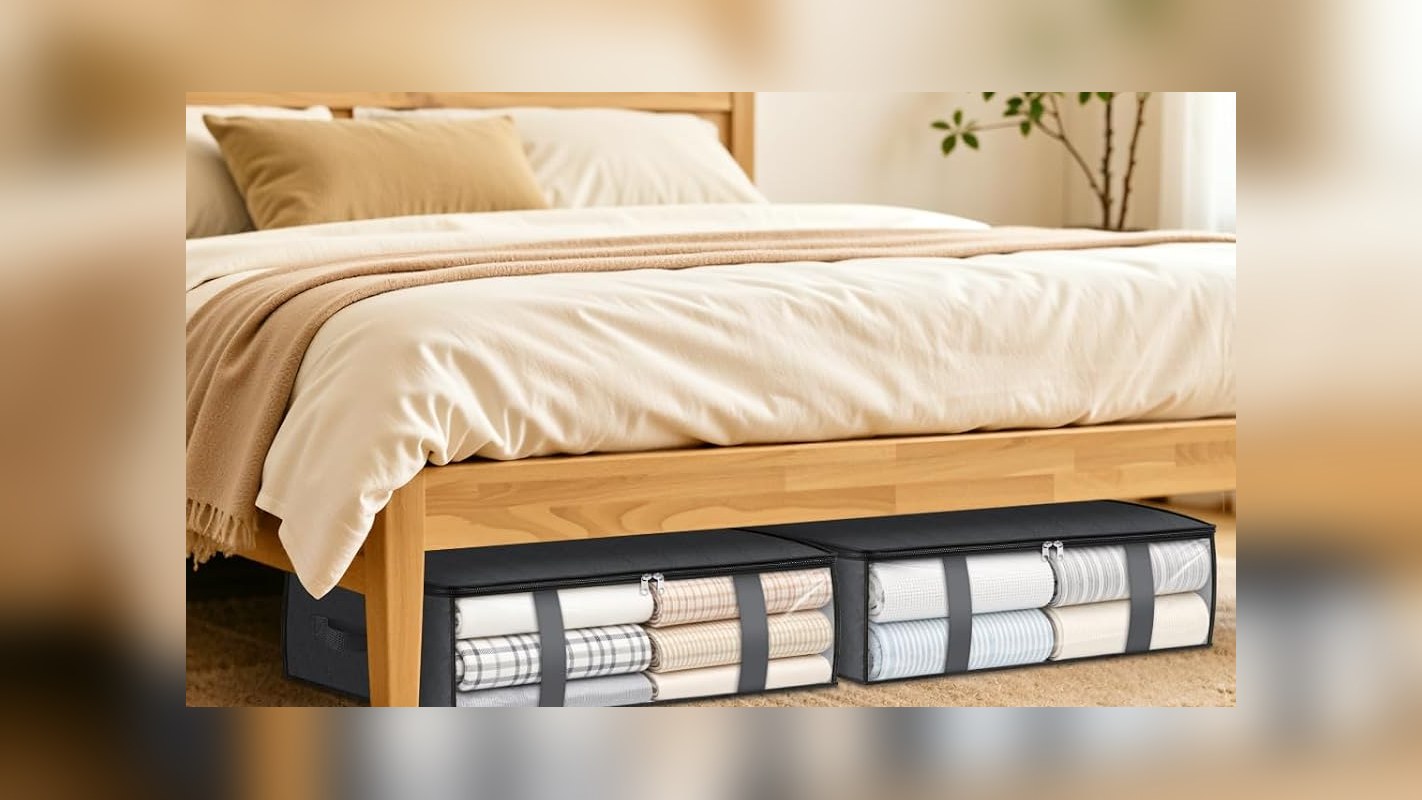 Two gray under-bed storage bags with clear windows holding folded blankets and sheets beneath a wooden bed frame